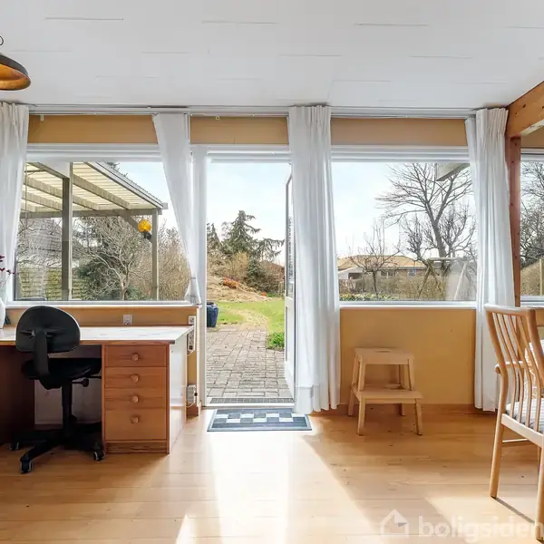 Et lyst rum med et skrivebord og stol til venstre, et spisebord og stole til højre. Store vinduer viser en have med fliser udenfor.