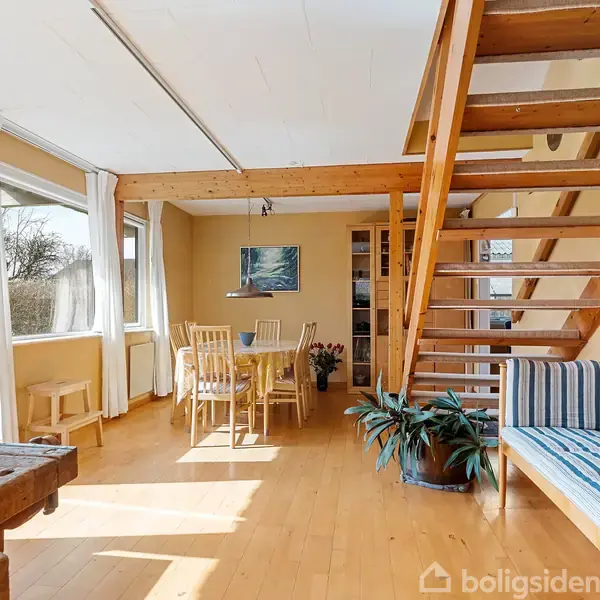 Trætrappe fører til anden etage i et lyst rum med trægulve. Spisebord og stole står ved store vinduer. Sofa og planter står under trappen ved en hvidvæg.