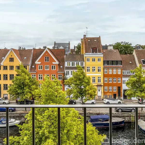 Farverige bygninger står langs en vej og kanal. Biler er parkeret foran husene, og både ligger i vandet. Træer med grønne blade pryder den hyggelige byscene.