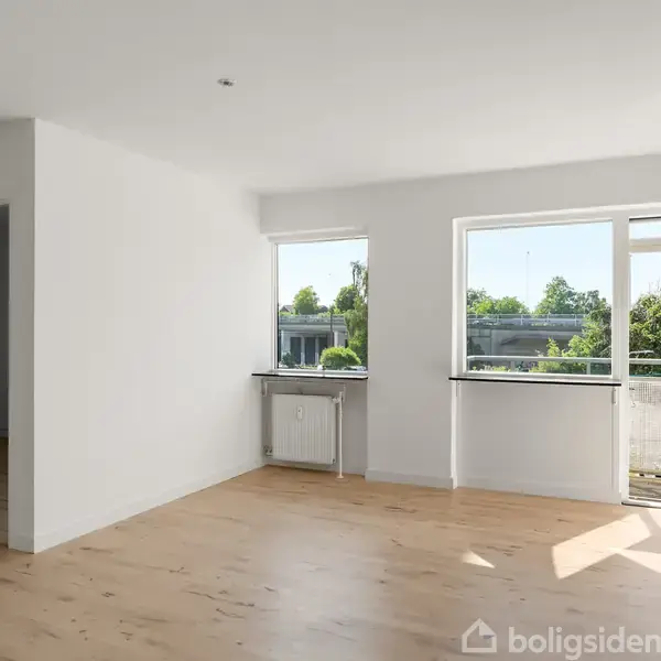 Et lyst, tomt rum med trægulv, hvor to vinduer og en glasdør åbner ud til en balkon. Udenfor ses træer og et hus i baggrunden. Ingen tekst er synlig.