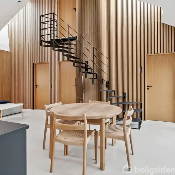 Rundt spisebord med fire stole står i et moderne stueområde med trægulv og minimalistisk design. En sort metaltrappe fører til en højere etage. Rummet er lyst og åbent.