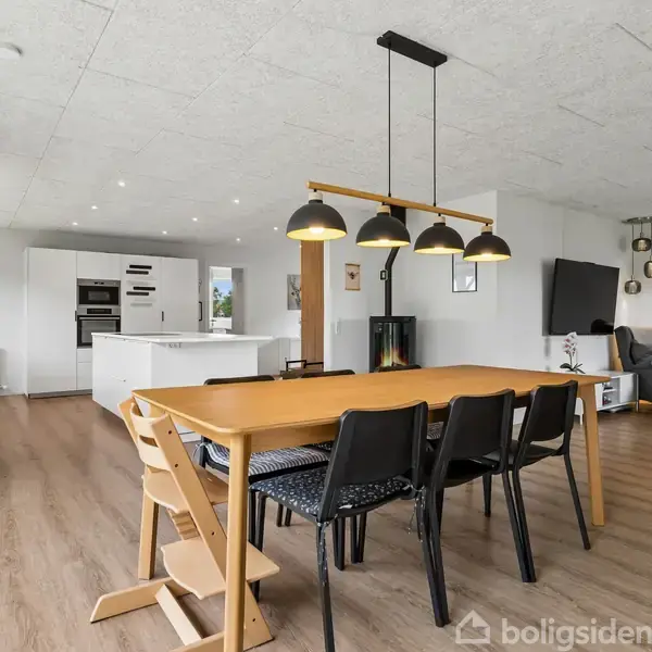 Spisebord med seks stole under hængende lamper i rummeligt, moderne køkken-alrum. I baggrunden ses hvidt køkken med ø og stueområde med sofa og vægmonteret TV.