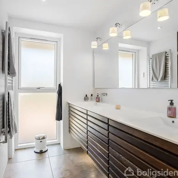 Badeværelse med dobbel håndvask i hvidt skab, lyst spejl og oplyst af lamper, med håndklæder på væggen og et vindue med sløret glas. Rummeligt og lyst design.