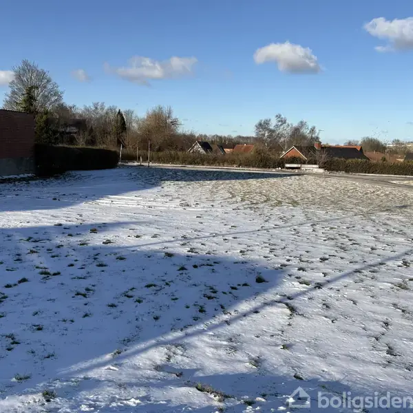 En snedækket mark strækker sig i en forstad med træer og huse i baggrunden. Skyer pryder en blå himmel, mens solen kaster skygger på sneen.