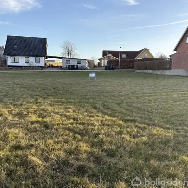 Græsplæne med et salgsskilt i midten står ubrugt. Et hvidt hus og en bil ses til venstre, mens to brune huse ligger til højre.