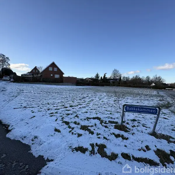En snedækket græsplæne med en blå vejskilt står til venstre for en vej, omgivet af huse i baggrunden. Skiltet tekst: "Bakkekammen".