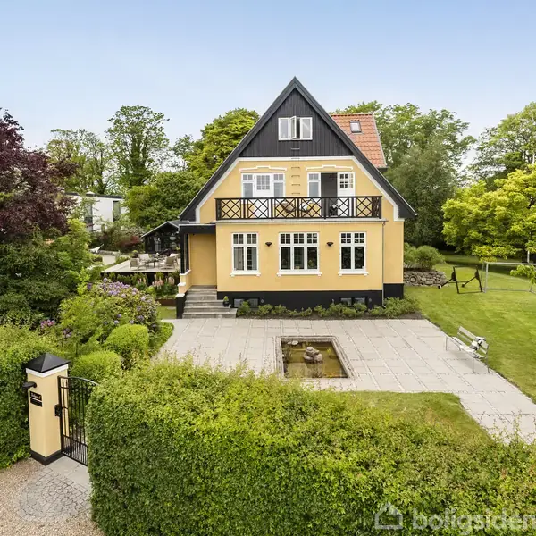 Et gult hus med sort tag står i en velplejet have. Der er en stenbelagt terrasse foran, omkranset af grønne hække og træer.