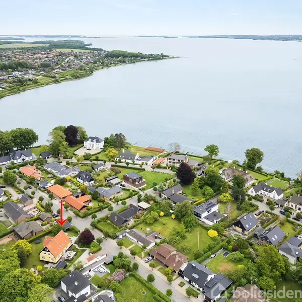 Hus med orange tag ses markeret med en rød pil blandt flere boliger i et grønt boligområde ved siden af en rolig sø.