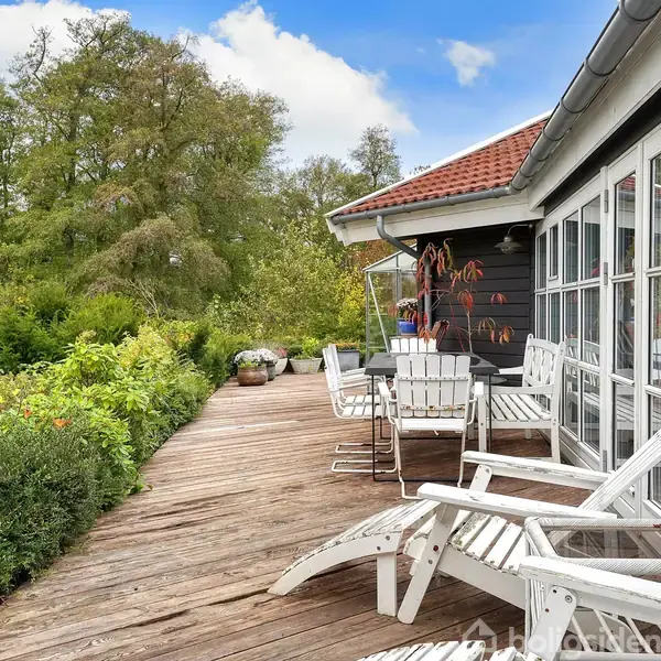 Havestol og bord står på en træterrasse, hvor de vender ud mod frodig natur. Store glasvinduer forbinder terrassen med et sortmalet hus med rødt tegltag i solskinsvejr.
