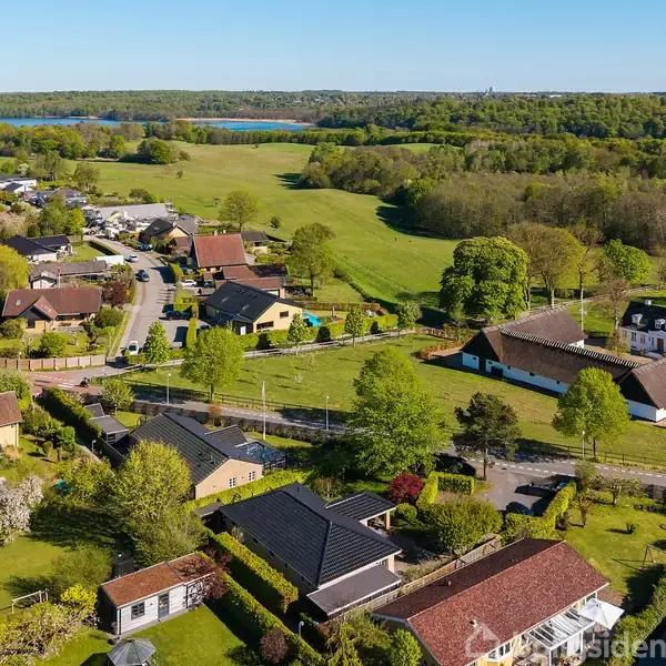 Luftfoto af et lille landsbysamfund med spredte huse omgivet af frodige grønne områder og marker. En bugtet vej snor sig gennem området, og der ses en sø i baggrunden.