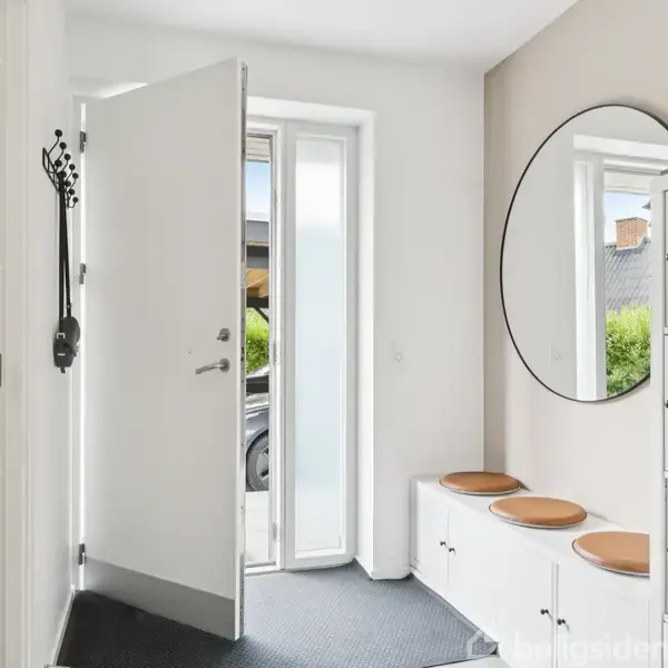 En indgang med en åben dør og et stort rundt spejl på væggen. Der er en bænk med skohylder og træpuder, samt en vaske- og tørremaskine station til venstre.