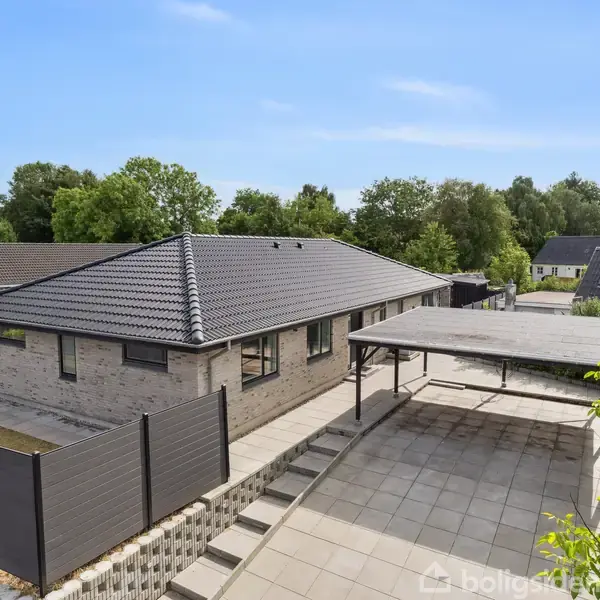 Et moderne, gråt murstenshus med sort tag står stille i et roligt villakvarter, omgivet af grønne træer og andre boliger. En flisebelagt indkørsel og carport er ved siden af huset.