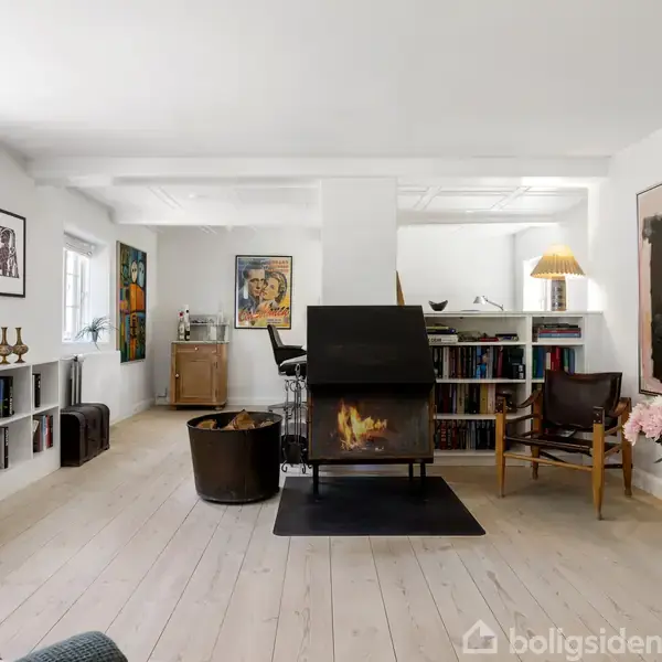 Pejs brænder i en lys stue med trægulve, omgivet af reoler og kunstværker på væggene. Der står lænestole omkring og en vase med pink blomster på gulvet.