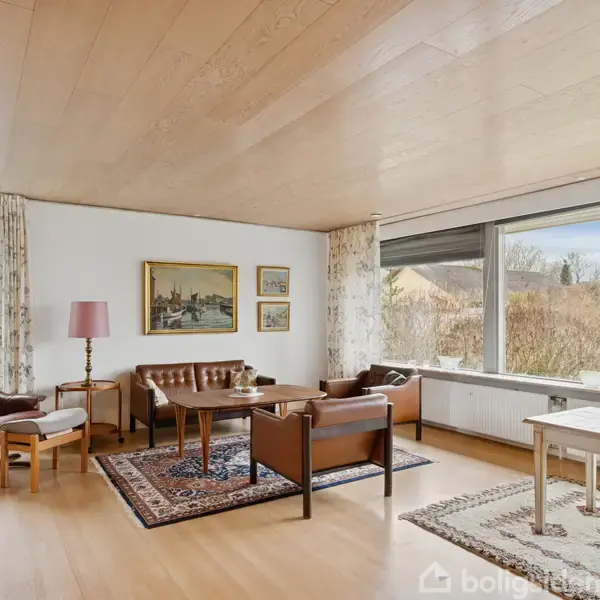 Stue med sofa og lænestol i læder, stilleben på tæpper. Store vinduer lader lys ind, afslører have. Træmøbler og malerier pryder væggen, skaber en hjemlig atmosfære.