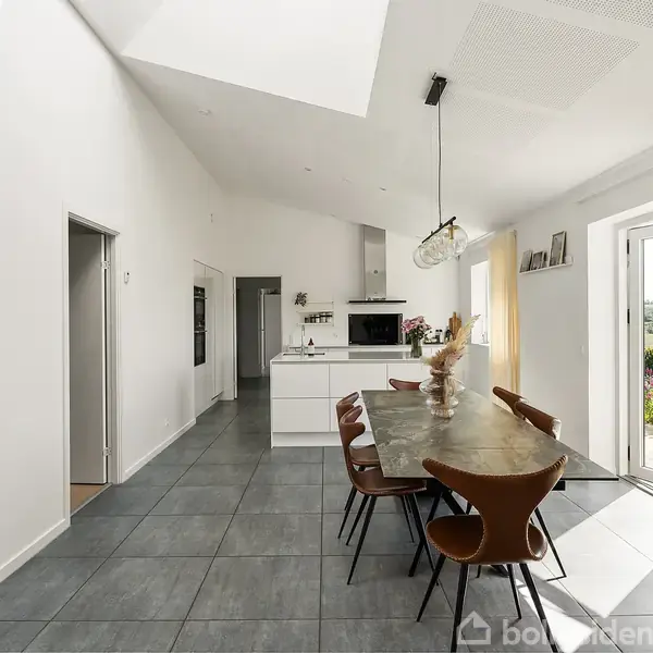 Et langt spisebord med brune stole omkring står i en moderne spisestue. Rummet har lyse vægge, store fliser på gulvet og glasdøre, der åbner mod en have med blomster.
