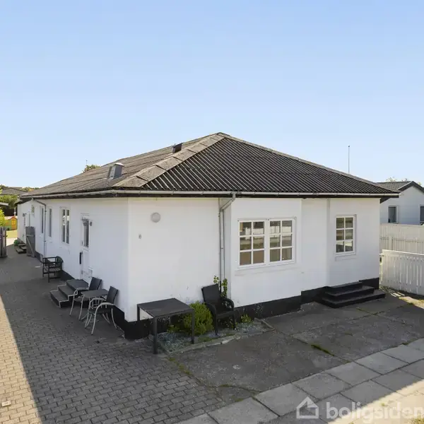 Hvid en-etagers bygning med sort tag, placeret på en asfalteret grund. Rundt omkring er der et hegn og flisebelægning. Flere stole står langs husets facade.