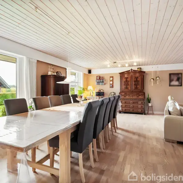 Langt spisebord med sorte stole står i en rummelig stue. Store vinduer slipper lys ind. Rummet har træloft, en sofa, skab og reoler i baggrunden.