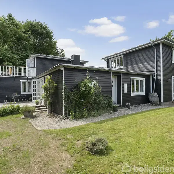 Et sortmalet hus med to etager og en terrasse har en velholdt græsplæne og jordsti omkring sig. Det er omgivet af grønne træer og buske under en skyfri himmel.