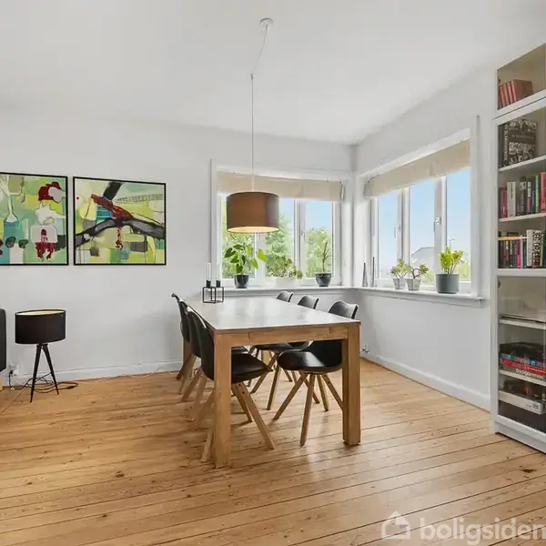 Spisebord med stole står i en lys spisestue med trægulv og en sort sofa. Store vinduer slipper lys ind, og en reol med bøger er placeret ved siden af kunst på væggen.