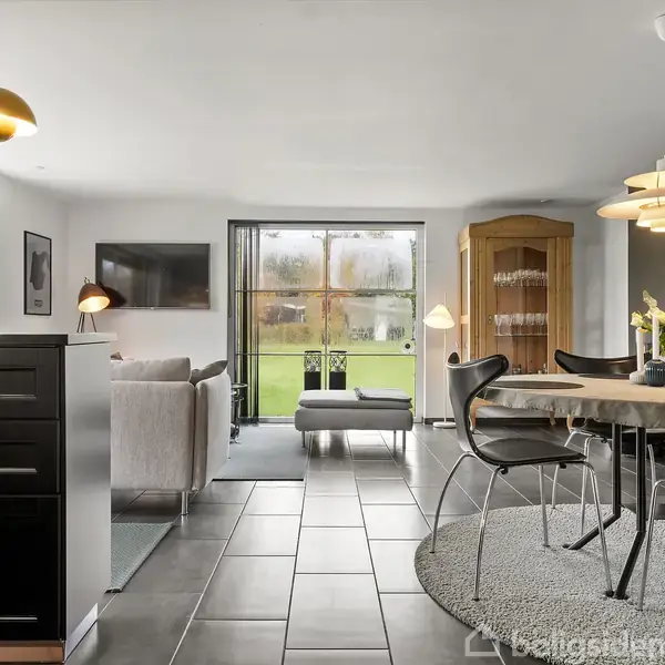 Et moderne køkken-alrum, hvor der ses et sort køkkenbord til venstre og en rund spisebord med stole til højre. Vinduer bagtil viser en grøn have. Lamper hænger over bordene.