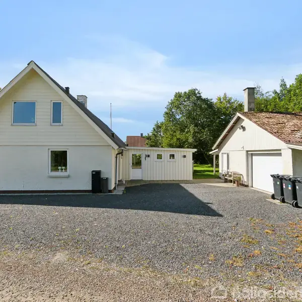 To hvidmalede bygninger står på en grusbelagt gårdsplads omgivet af frodigt grønt. Den ene har et skråt tag med kvistvinduer, mens den anden har tag med mos og skraldespande foran.