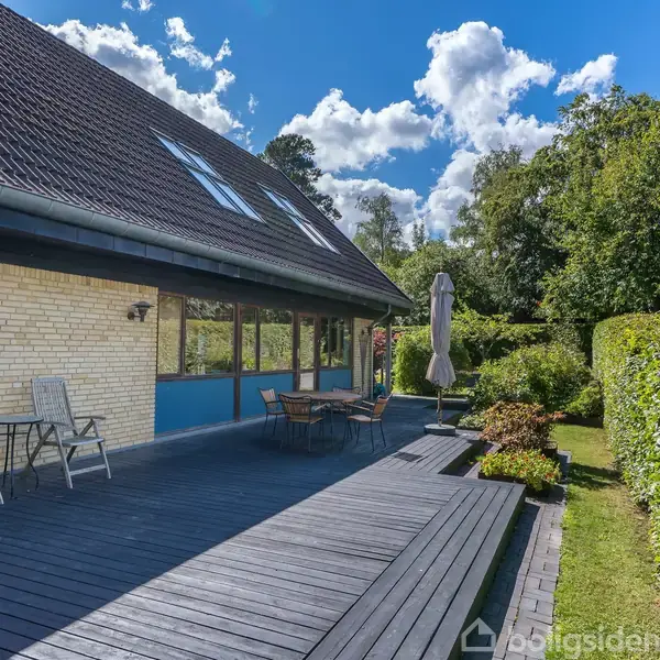 Et stort udendørs trædæk med borde og stole, omgivet af en velplejet have med buske og træer. Huset har et sort tegltag og store vinduer, under en klar blå himmel.