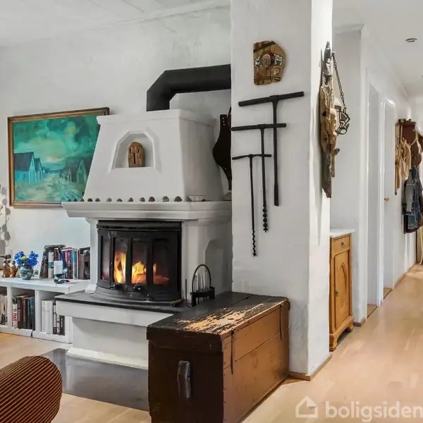 En charmerende pejs brænder i stuen, omkranset af hylder med bøger og dekorationer. Ved siden af pejsen står en brun kiste. En lang gang fører videre ind i huset.