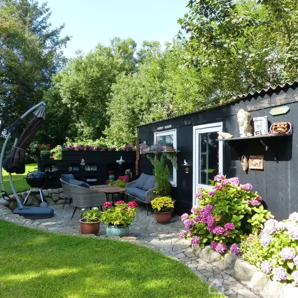 En sortmalet bygning med udendørs siddeområde, omgivet af blomster og planter i farverige krukker, står på en stenterrasse i en grøn have med træer i baggrunden.
