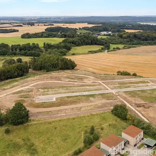 Luftfoto viser åbent landskab med marker og skov. En vej er under konstruktion i forgrunden nær nogle få huse. Det omkringliggende område er landligt med åbne marker.