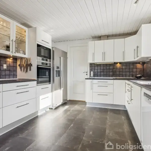 Et moderne køkken med hvide skabe og sorte fliser har indbyggede ovne, køleskab og vask. Knivholder på væggen, skuffer åbne, og vindue til højre lukker lys ind.