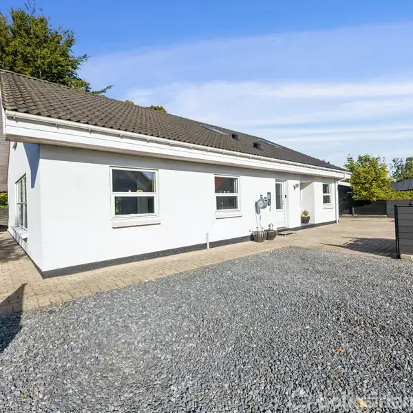 Et hvidt enfamiliehus med skråt tag står på en grusbelagt indkørsel. Huset er omgivet af en terrasse og træer under en klar blå himmel.