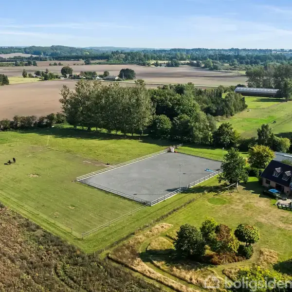 Et landsted med en stor ridebane ligger i en frodig, grøn mark. Nogle træer omgiver området, og der er en mindre bygning ved siden af ridebanen. Tekst: "EDC".