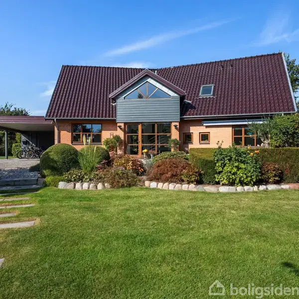 Et moderne hus med rødt tegltag står omgivet af en velplejet have med buske og græsplæne. Der er en stenbelagt sti foran huset og en carport til venstre.