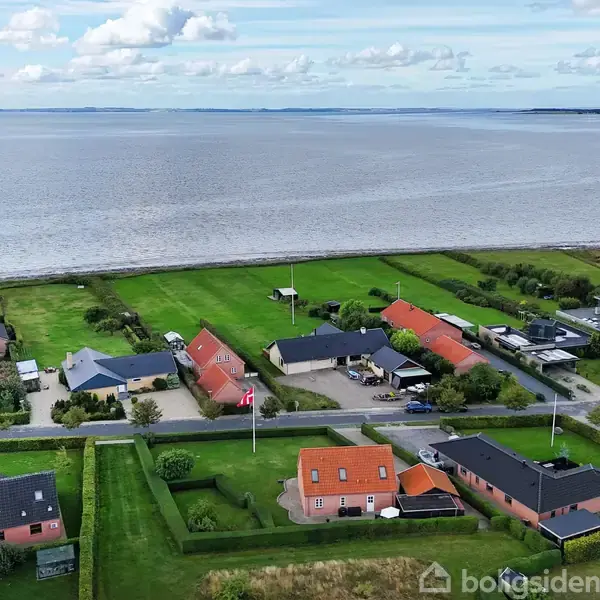 Huse ligger langs en vej med haver, flagstænger og grønne marker. Havet strækker sig i baggrunden under en let overskyet himmel.