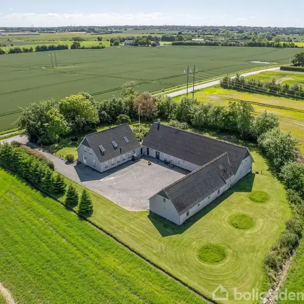 En gård med to langstrakte, hvide bygninger står omkring en gårdsplads. Bygningerne er omgivet af grønne marker under en åben himmel i landlige omgivelser.