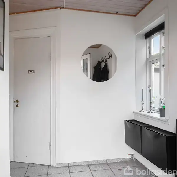 Et hvidt indgangsområde med klinkegulv, en lukket dør til venstre mærket "WC", en rund spejling over for, en vindue til højre med stearinlys på vindueskarmen og sorte opbevaringsenheder nedenunder.