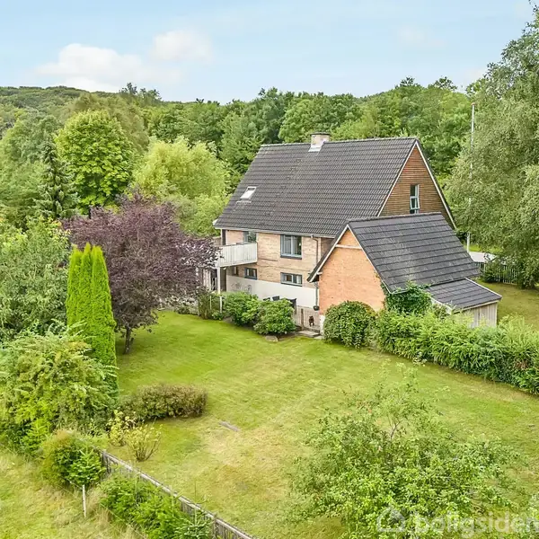En to-etagers murstensvilla med skråt tag står stille omgivet af en frodig have med græsplæne, buske og træer. I baggrunden ses skovklædte bakker under en blå himmel.