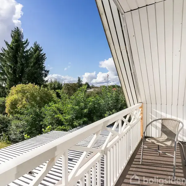 En balkon med en stol står på et trædæk; omgivet af en grøn have og store træer under en blå himmel med hvide skyer.