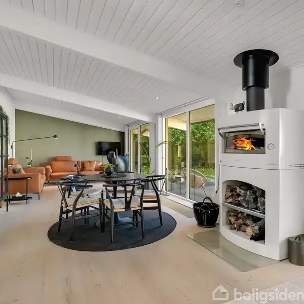 Pejs brænder i stue med hvide vægge og loft. Rundt, sort spisebord med fire stole står på tæppe. Store vinduer giver udsigt til grønne omgivelser. Ordet "Boligsiden" ses nederst.