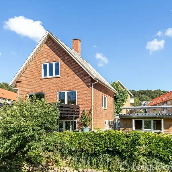 Rødt murstenshus med spidst tag står stille omgivet af grønt løv og buske i et boligområde med solskin og blå himmel. Andrse huse ses i baggrunden.