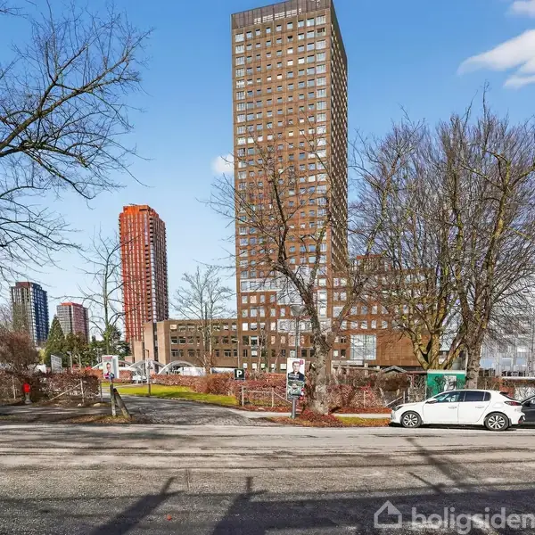 En høj bygning står blandt flere træer i et urbant område, med flere biler parkeret langs vejen. Andre mindre bygninger og et højhus ses i baggrunden under en blå himmel.