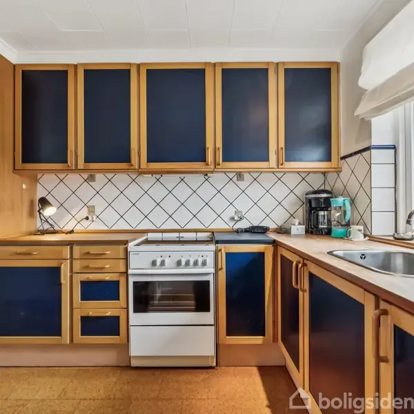 Et køkken med blå skabe og skuffer i trærammer. En hvid komfur står i midten, omgivet af en kaffeautomat og vask. Vindue med udsigt til himmel og bygning.