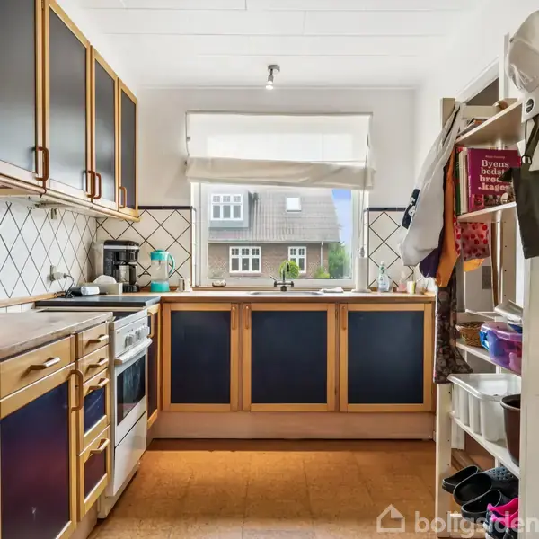 En køkken med træskabe og mørke paneler står ved et vindue. Køkkenet har en ovn, vask og åbne hylder med forskellige genstande. Udenfor er der udsigt til et traditionelt hus.