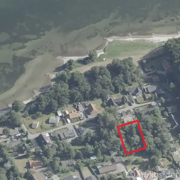 Et landområde markeret med en rød firkant ligger i et villakvarter nær kysten og grønne skove. Området ses fra oven med tydelige huse og veje.