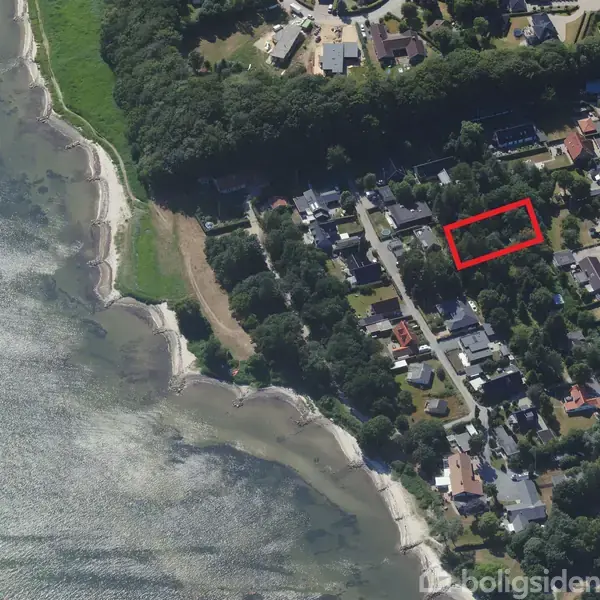 Luftfoto viser et kystområde med boliger nær en strand. En rød rektangel markerer en grund blandt husene. Omgivelserne er præget af træer og tætliggende bebyggelse.