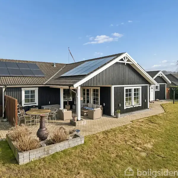 Sortmalet hus med solpaneler på taget står stille i en frodig have, omgivet af havemøbler og en blå himmel.