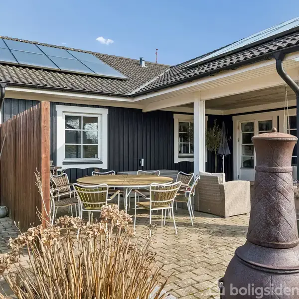 Et udendørs terrasseområde med møbler, som inkluderer et rundbord og stole, i en have ved et moderne hus med solpaneler på taget. En havepejs står i forgrunden.