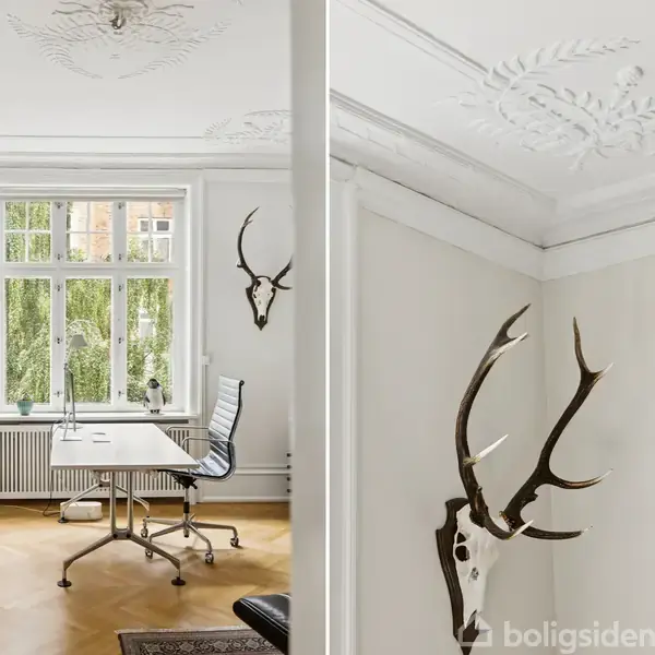 Et kontor med et skrivebord og en kontorstol placeret foran et stort vindue. Væggene er dekoreret med hjortegevirer, og loftet har detaljerede stukdekorationer.