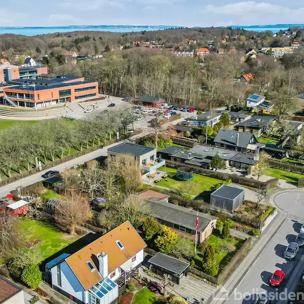 En beboelsesområde med huse, nogle med orange tage, omkranset af træer og en græsplæne ved siden af et større moderne bygningskompleks med parkeringsplads, set fra luften.