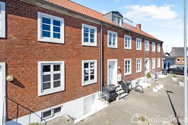 Rækkehus i røde mursten med hvide vinduer og døre står stille på en brostensbelagt gade. En cykel og skraldespand er ved siden af hoveddøren.